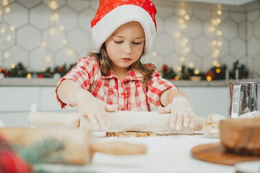 Little dark-haired girl 3 years old in red Christmas cap and checkered shirt roll out dough for gingerbread cookies in white Christmas decorated kitchen with garland lights. Merry Christmas, New Year