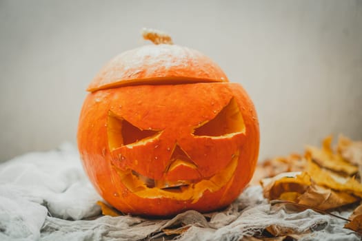 Orange bright halloween pumpkin with carved nose, mouth and eyes on light gray background next to autumn yellow leaves. Scary face carved on vegetable. Halloween holiday decoration