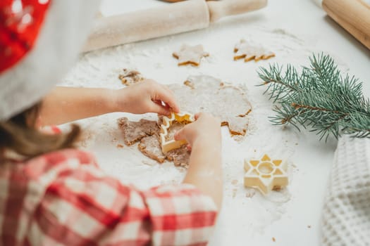 Partially blurred hands of little girl cut out Christmas tree gingerbread cookies from rolled dough in white kitchen. Child prepares cookies on white table strewn with flour