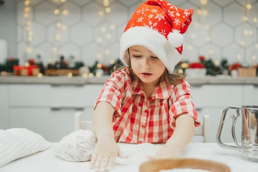Little dark-haired girl 3 years old in red Christmas cap and checkered shirt prepares dough for gingerbread cookies in white Christmas decorated kitchen with garland lights. Merry Christmas, New Year