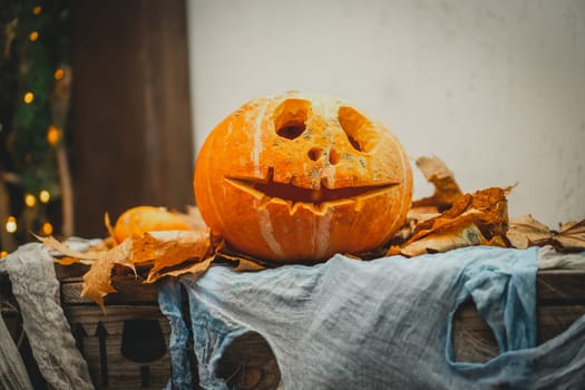 Orange bright halloween pumpkin with carved nose, mouth and eyes on wooden box with gauze as web next to autumn yellow leaves. Scary face carved on vegetable. Halloween holiday decoration