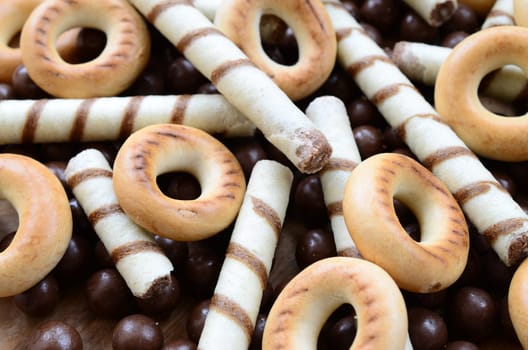 A lot of crispy sweet tubules, chocolate melting balls and yellow bagels lie on a wooden surface. Close up view