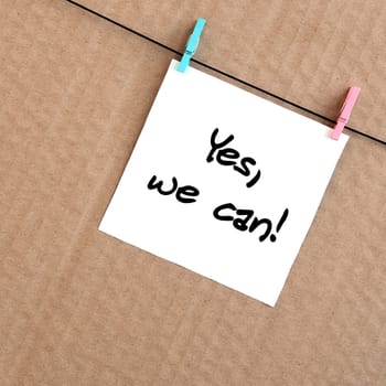 Yes, we can! Note is written on a white sticker that hangs with a clothespin on a rope on a background of brown cardboard