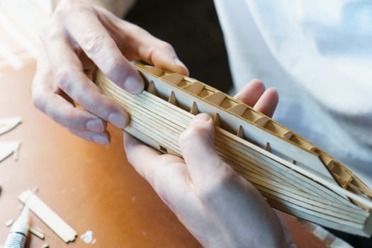 Hands of man gluing plywood details for ship model with glue, holding with fingers. Process of building toy ship, hobby, handicraft. Table with various materials, parts and devices for work