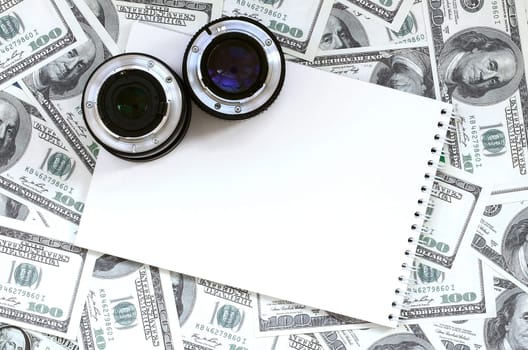Two photographic lenses and white notebook lie on the background of a lot of dollar bills. Space for text