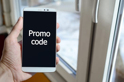 A person sees a white inscription on a black smartphone display that holds in his hand. Promo code