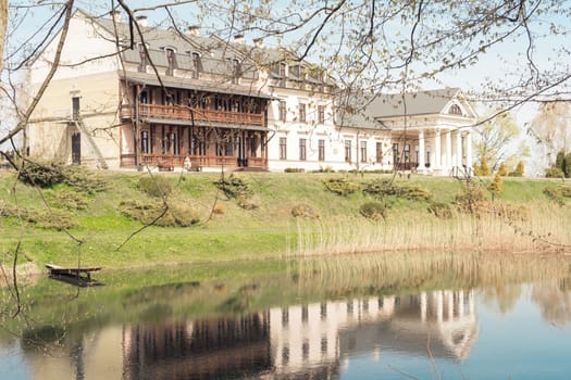 Belarus, village Radzivilki, 01.05.22. Restored old manor Svyatsk Gkursky in Belarus. View to main entrance with columns on bank of small pond with reeds. House is reflected in water