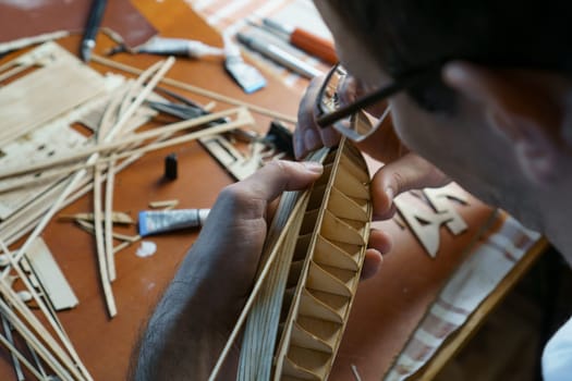 Hands of man gluing plywood details for ship model with glue, holding with fingers. Process of building toy ship, hobby, handicraft. Table with various materials, parts and devices for work