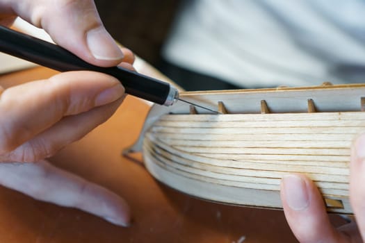 Hands of man removing excess glue from plywood details for ship model with utility knife. Process of building toy ship, hobby, handicraft. Table with various materials