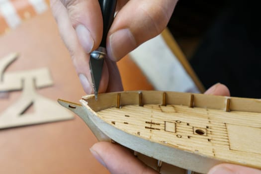 Hands of man cutting out details for ship model with clerical knife from plywood. Process of building toy ship, hobby and handicraft. Table with various materials, parts and devices for work
