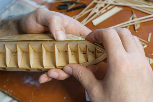 Hands of man gluing plywood details for ship model with glue, holding with fingers. Process of building toy ship, hobby, handicraft. Table with various materials, parts and devices for work