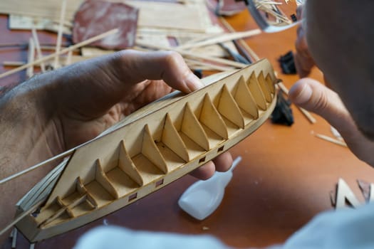 Hands of man gluing plywood details for ship model with glue, holding with fingers. Process of building toy ship, hobby, handicraft. Table with various materials, parts and devices for work