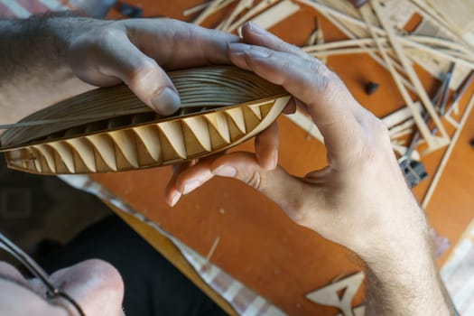 Hands of man gluing plywood details for ship model with glue, holding with fingers. Process of building toy ship, hobby, handicraft. Table with various materials, parts and devices for work