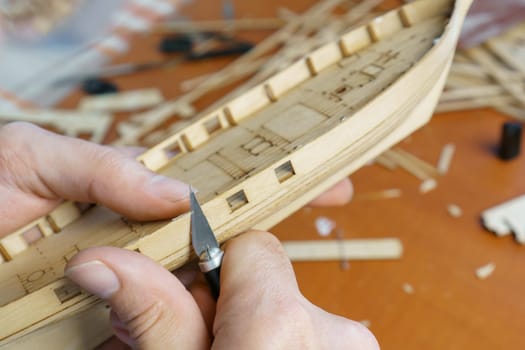 Hands of man cutting out details for ship model with clerical knife from plywood. Process of building toy ship, hobby and handicraft. Table with various materials, parts and devices for work