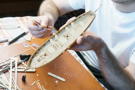 Hands of man adjusts plywood details for ship model, grinding on sandpaper. Process of building toy ship, hobby and handicraft. Table with various materials, parts and devices for work
