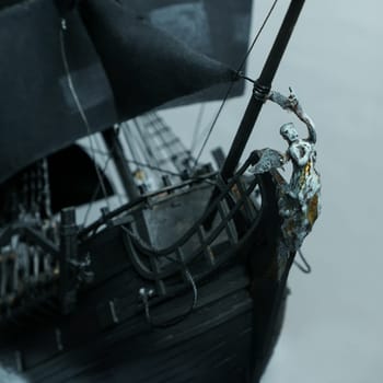 Pirate ship model Black pearl made of plywood, with miniature figures of pirate sailors, close-up details. Bow with figure of woman with wings. Ship sails on waves with patched sails