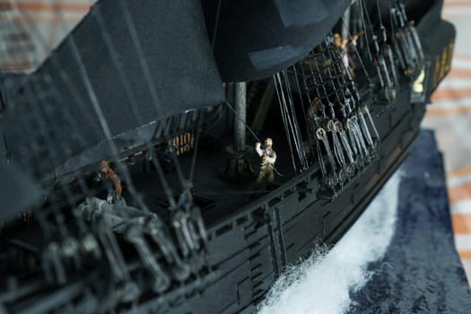 Pirate ship model Black pearl made of plywood, with miniature figures of pirate sailors, close-up details. Ship sails on wave. Sailors on deck pulling ropes, partially blurred. Patched sails belly out