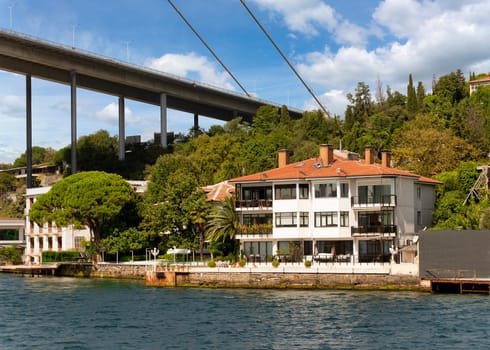 View from the sea of the European side of Bosphorus strait, Istanbul, Turkey, with traditional waterfront house, under Bosphorus bridge, and green hill with dense trees, in a summer day