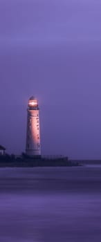 A lighthouse on the coast of the sea at dusk, its bright lamp is a landmark for ships. A lighthouse in the lilac twilight. Seascape, nature background.