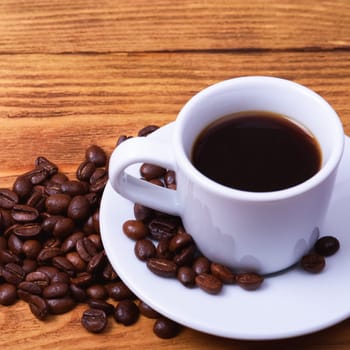 Freshly made coffee in a white Cup on a white saucer surrounded by scattered coffee beans on a wooden table. Fresh roasted coffee. Place for an inscription or logo.