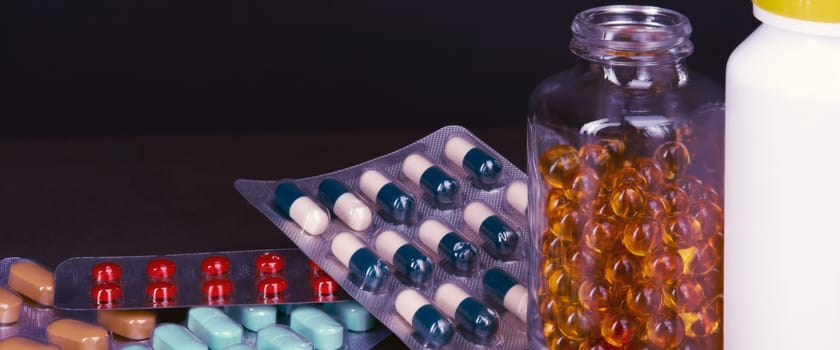Pills and tablets of different colors are scattered and in bottles on a dark background