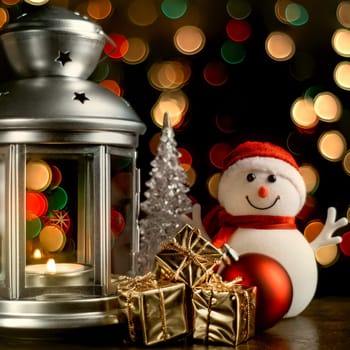 Christmas composition: A snowman, a lantern with candles, bright shiny boxes with gifts, blurred bokeh lights in the background.