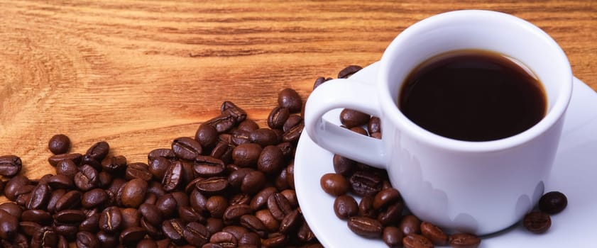 Freshly made coffee in a white Cup on a white saucer surrounded by scattered coffee beans on a wooden table. Fresh roasted coffee. Place for an inscription or logo.