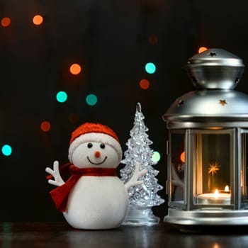 Christmas composition: a snowman, a lantern with candles and blurred bokeh lights in the background.