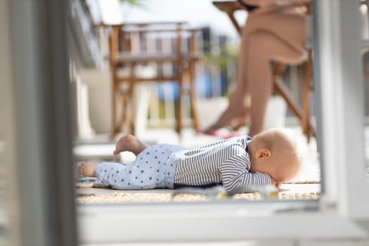 Cute little infant baby boy sulking while playing with toys outdoors at the patio in summer being supervised by her mother seen in the background