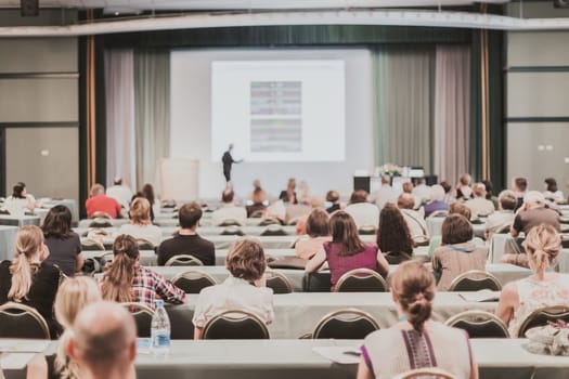 Business and entrepreneurship symposium. Speaker giving a talk at business meeting. Audience in the conference hall. Rear view of unrecognized participant in audience.