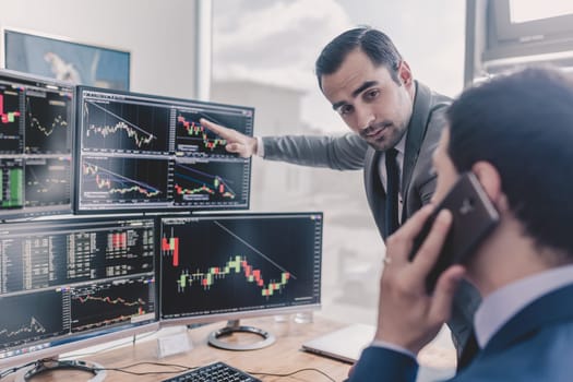 Businessmen trading stocks online. Stock brokers looking at graphs, indexes and numbers on multiple computer screens. Colleagues in discussion in traders office. Business success concept.