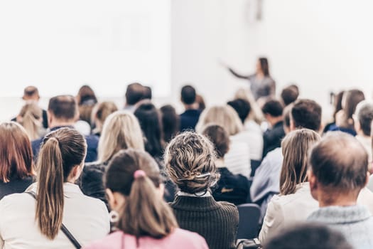 Business and entrepreneurship symposium. Female speaker giving a talk at business meeting. Audience in conference hall. Rear view of unrecognized participant in audience.