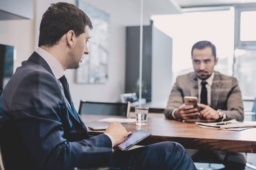 Image of two young businessmen using smart phones and touchpad at meeting in moder office.