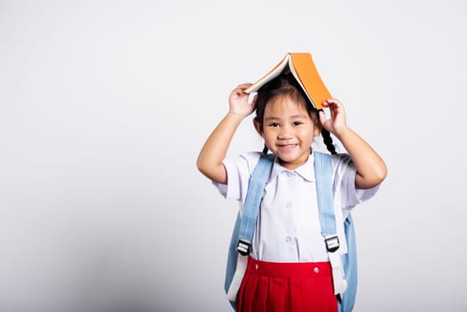 Asian adorable toddler smiling happy wearing student thai uniform red skirt stand holding book over head like roof in studio shot isolated on white background, Portrait little children girl preschool