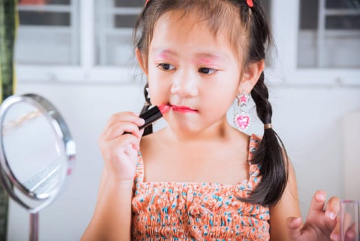 Asian adorable funny little girl making makeup her face she look in the mirror and applying red lipstick to mouth, Learning activity to be woman, happy kid is beautiful make up with cosmetics toy