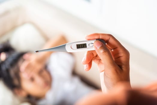 Sick kid. Mother checking temperature of her sick daughter with thermometer in mouth, child laying in bed taking measuring her temperature for fever and illness