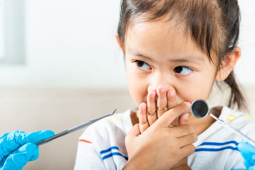 Dental kid examination. Doctor examines oral cavity of child uses mouth mirror to check teeth cavity but little girl cover mouth no need to checking she afraid, scared and closes his mouth with hands