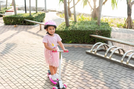 Child riding scooter. Happy Asian little kid girl wear safe helmet playing pink kick board on road in park outdoors on summer day, Active children games outside, Kids sport healthy lifestyle concept