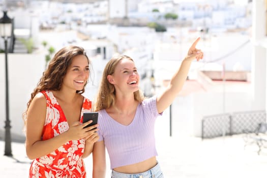 Two happy tourists checking smart phone pointing landmark standing in a white town of andalusia