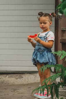 Concentrated little girl 3-4 years old with two ponytails eats watermelon. Child outdoors against background of building holds sweet healthy fruit in her hands. Summer, harvest, vitamins