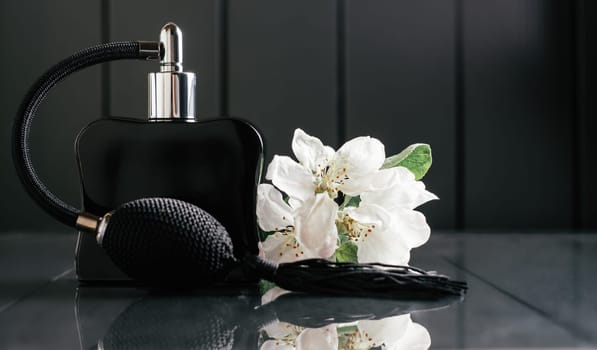 Black bottle of eau de toilette or perfume with long tassel spray pomp stands on dark background on reflective surface with apple blossom. Copy space