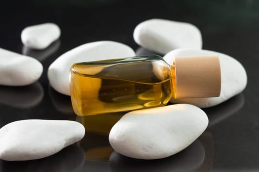 Transparent glass bottle of yellow oil or perfume, lying on white stones on a black reflective surface