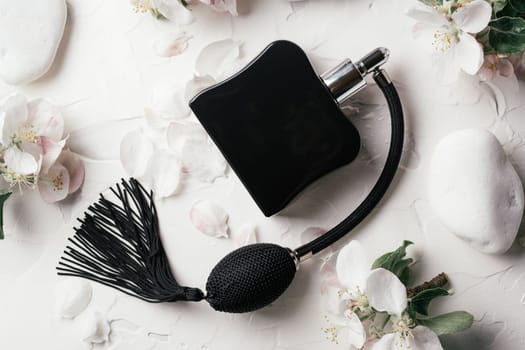 Black bottle eau de toilette or perfume with long tassel spray pomp on delicate background of apple flowers, petals and pebble stones lying on white gypsum plaster. Top view