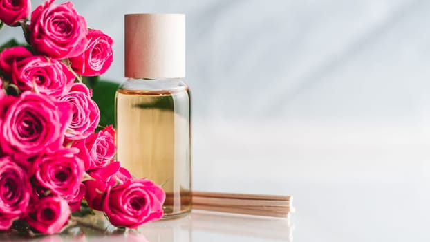 Clear bottle of yellow oil or perfume with wooden sticks and delicate pink roses on white surface. SPA salon and procedures concept. Banner. Copy space