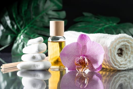 Beautiful lilac orchid flower, clear bottle of yellow oil or perfume, wooden sticks and rolled towel with stack of white stones and monstera leaves, on black surface. Spa salon and procedures concept