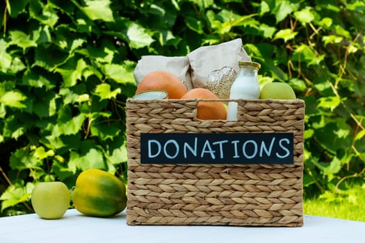 Cereals, canned food, milk, vegetables and fruits in a box for food donations on a white table on a background of greens. Charity and donation concept, copy space.