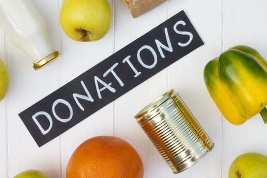 Cereals, canned food, milk, vegetables and fruits with a chalk Donations sign on a white wooden table. Flat lay. Charity and donation concept.