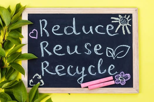 Chalk board Reduse Reuse Recycle sign on a yellow background with green leaves and chalk pieces. Top view
