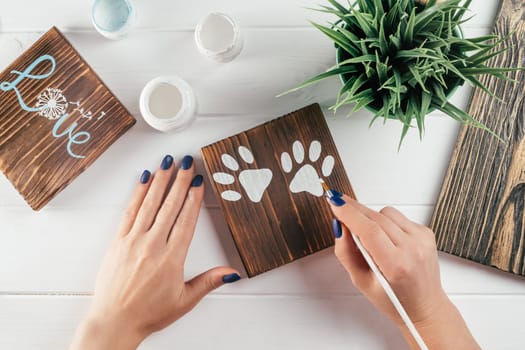 Womans hand draws paw prints on decorative board made of burnt wood with brush of white paint. Creative DIY