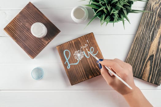 Womans hand draws word Love and dandelion flower on decorative board made of burnt wood with brush of blue paint. Creative DIY